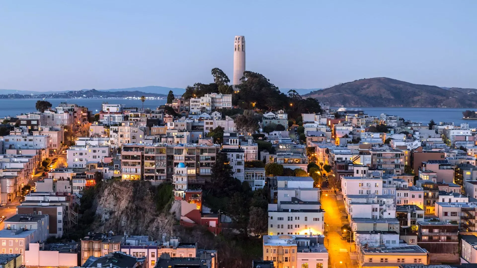 황혼 무렵의 샌프란시스코 Coit Tower 타워 앞에는 조명이 켜진 거리가 있고, 타워 뒤로는 샌프란시스코 만이 보입니다.