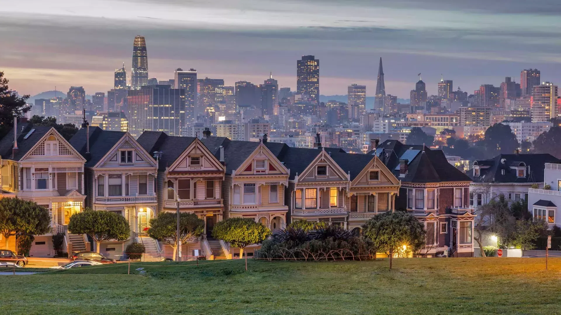 San Francisco's Painted Ladies, a row of Victorian houses, are pictured against a backdrop of the city skyline at twilight.